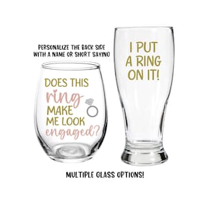 May include: Two clear glass cups with gold and pink text. The wine glass says "Does this ring make me look engaged?" and the beer glass says "I put a ring on it!"