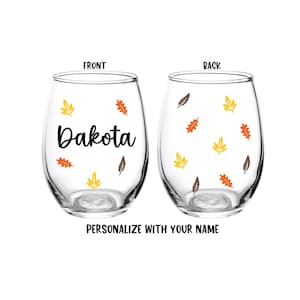 May include: Two clear stemless wine glasses featuring a pattern of small yellow, orange, and brown autumn leaves. The glass on the left displays the name Dakota in black script. The text FRONT, BACK, and PERSONALIZE WITH YOUR NAME are visible.