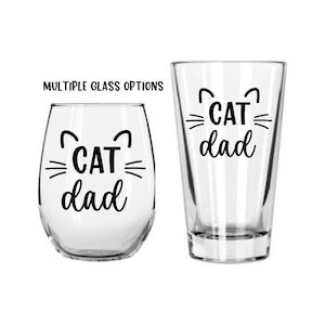 May include: Two clear glass cups with black text that reads "CAT dad" and a stylized cat face. The glass on the left is a wine glass and the glass on the right is a pint glass.