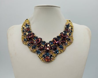 May include: A gold statement necklace with large, faceted, dark red, blue, and pink gemstones. The necklace is layered with smaller gold beads and gemstones.