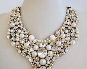 May include: A statement necklace featuring a bib design. The necklace is densely adorned with a mix of faux pearls, clear crystals, and gold-toned accents. The crystals vary in size and shape, creating a sparkling effect. The necklace is displayed on a white mannequin.