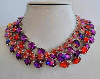 Peut inclure: Un collier ras de cou à plusieurs rangs avec un dégradé de cristaux orange, rouge et violet. Le collier est fait de métal argenté.