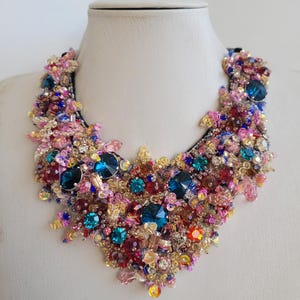 Puede incluir: Un llamativo collar con una gran cantidad de gemas y cuentas de colores. El collar muestra una vibrante mezcla de azules, rosas y dorados, creando un diseño lujoso y llamativo. Los intrincados detalles y las variadas texturas lo convierten en una pieza destacada.