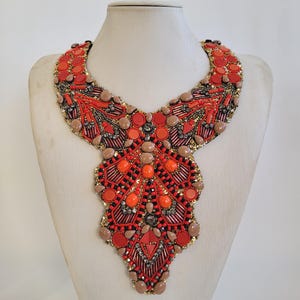 May include: A statement bib necklace in a striking design. The necklace is adorned with red, orange, and brown beads, sequins, and stones. The necklace is displayed on a mannequin.