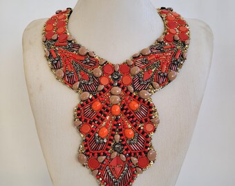 May include: A bib necklace with a vibrant design. The necklace features a mix of red, orange, and brown beads, sequins, and stones. The necklace is displayed on a mannequin.