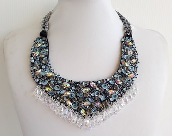 May include: A statement necklace with a dark blue beaded base, adorned with iridescent crystals, light blue beads, and clear crystal teardrops. The necklace has a silver chain and is displayed on a white mannequin.