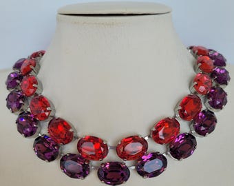 Peut inclure: Un collier ras de cou à deux rangs avec de gros strass ovales rouges et violets. Le collier est en métal argenté.