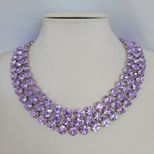May include: A silver necklace with three rows of light purple rhinestones. The necklace is on a white mannequin bust.