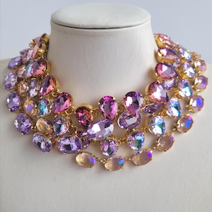 Puede incluir: Collar llamativo de varias capas con grandes cristales iridiscentes en forma de lágrima y ovalados en tonos morados, rosas y melocotón. Los cristales están engarzados en un marco de metal dorado, creando un diseño glamuroso.