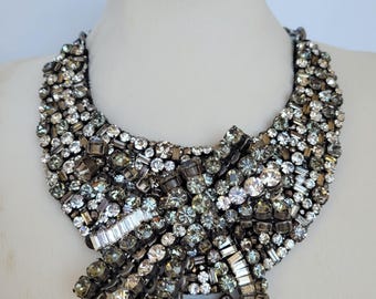 May include: A statement necklace with a bib design, densely covered in various sizes and shapes of sparkling crystals. The crystals are primarily silver, gray, and clear, creating a glamorous and eye-catching accessory.
