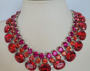 May include: A multi-layered statement necklace with large, oval-shaped red, orange, and pink rhinestones. The necklace is made of silver-toned metal.