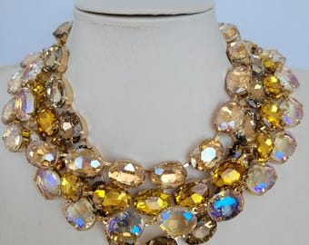 May include: A gold tone statement necklace with multiple rows of large, oval-shaped, iridescent crystals in shades of amber and champagne.