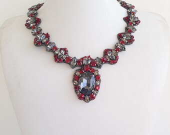 Peut inclure: Un collier statement avec une grande pierre ovale gris foncé entourée de strass rouges. Le collier est en métal argenté et présente un design délicat et ondulé.