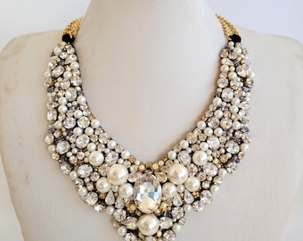 May include: A statement necklace featuring a bib design. The necklace is adorned with a mix of white pearls, clear crystals, and a large, central, faceted gemstone. The necklace has a gold-tone chain and black fabric at the clasp.