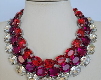 May include: A multi-layered statement necklace with large oval-shaped rhinestones. The necklace features three rows of rhinestones in shades of red, pink, and clear.