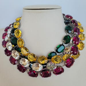 May include: A multi-layered statement necklace featuring large oval-shaped crystals in shades of red, yellow, green, and clear. The necklace is made of metal and has a silver finish.