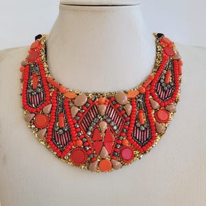 May include: A striking bib necklace in shades of red, orange, and tan, with gold-coloured detailing. The necklace is adorned with various beads and stones, creating a textured effect. Displayed on a mannequin.