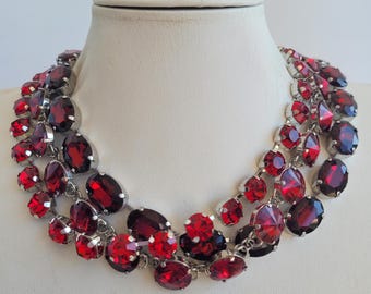 May include: A multi-layered statement necklace with large, red, faceted crystals. The necklace is made of silver-toned metal and features a variety of crystal shapes and sizes.