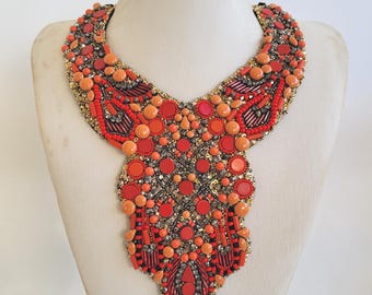 May include: A vibrant bib necklace with a striking design. The necklace features a mix of red, orange, and gold beads and embellishments. The design includes circular and rectangular elements, creating a bold and eye-catching accessory.