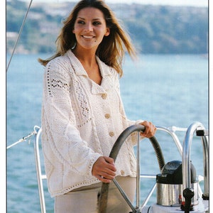 Puede incluir: Una mujer con un cárdigan blanco de punto con un patrón de punto de cable y botones, de pie en un barco y sujetando el volante.