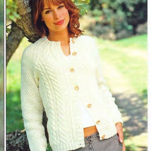 May include: A white cable knit cardigan with a button closure. The cardigan is worn by a person with red hair and is photographed outdoors.