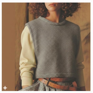 May include: A grey knitted vest with a crew neck and ribbed hem. The vest is worn over a cream-coloured long-sleeved shirt. A brown leather belt with a square buckle is visible. The style is casual and comfortable.