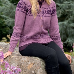 May include: A purple cardigan with a snowflake pattern and button fastening. The cardigan is being worn by a person with long blonde hair and black trousers. The person is sitting on a rock with purple flowers in the foreground.