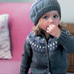 May include: A young child wearing a gray knitted cardigan with a white and purple patterned design. The cardigan has a button closure and the child is wearing a gray knitted hat with a white pom-pom on top.