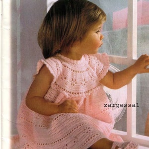 May include: A pink crocheted baby dress with a scalloped neckline and short sleeves. The dress is worn by a baby girl who is looking out a window. The text "zargessal" is visible in the lower right corner of the image.