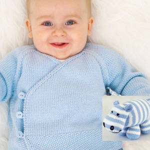 May include: A baby wearing a light blue knitted wrap sweater with three buttons. A knitted blue and white striped hippo toy is next to the baby. The baby is lying on a white fluffy surface.