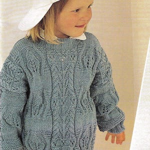 May include: A light blue knitted jumper with a cable knit pattern. The jumper has a round neckline and long sleeves. A child is wearing the jumper and a white baseball cap.