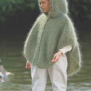 Op de afbeelding: Een jong persoon draagt een lichtgroene poncho met capuchon en een pluche textuur. De poncho is gebreid en heeft een lang, vloeiend ontwerp. De persoon staat in de buurt van een waterpartij.