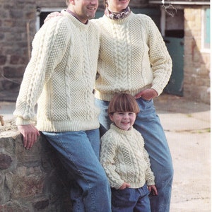 Pode incluir: Uma família de três pessoas vestindo suéteres de tricô de cabo cor creme. Os suéteres têm um design irlandês tradicional. A família está em pé em frente a uma parede de pedra.