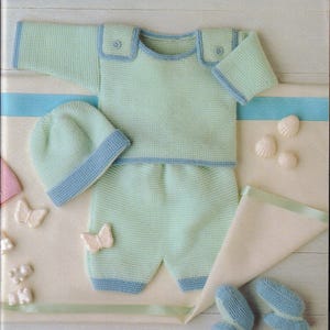 May include: A knitted baby outfit in light green with blue trim, including a sweater, shorts, hat, and booties. The set is decorated with small butterfly accents and seashells. The outfit is displayed on a white and cream surface.