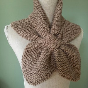 May include: A hand-knitted, taupe-colored scarf displayed on a mannequin. The scarf features a unique design with a knotted center and wide, flared ends. The chunky knit texture is visible throughout the scarf, showcasing the craftsmanship.