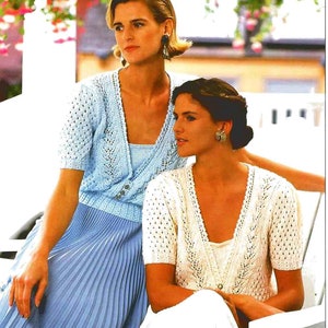 May include: Two women in knitted outfits. One wears a light blue short-sleeved cardigan with a matching top and a pleated skirt. The other wears a cream-coloured short-sleeved cardigan and a white top. The outfits have a delicate, openwork design.