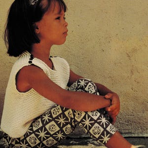 May include: A young person wearing a white knitted vest with dark buttons, patterned pants, and a headband. The pants feature a repeating geometric design in brown and white. The person is sitting with their legs crossed, and shoes are visible.