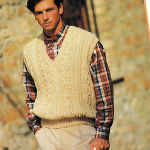 May include: A beige cable-knit vest with a V-neck, worn over a red, blue, and white plaid shirt. The vest has a textured pattern and is paired with beige corduroy pants. The person is holding a white object.