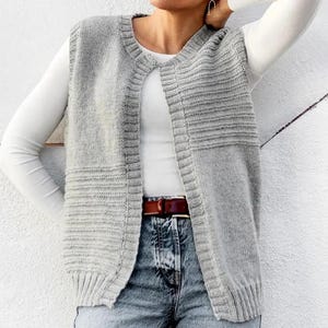Peut inclure: Un gilet en tricot gris clair avec un motif côtelé. Le gilet sans manches est ouvert sur le devant. Le modèle porte un haut blanc à manches longues, un jean bleu et une ceinture marron.