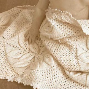 Könnte beinhalten: Eine cremefarbene gehäkelte Babydecke mit einem zarten Blumenmuster. Die Decke ist aus weichem Garn gefertigt und hat eine zarte Spitzenborte.