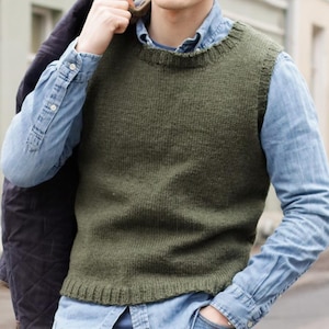 May include: An olive green knitted vest with a ribbed hem and neckline. The vest is worn over a light blue denim shirt. A dark navy jacket is draped over the shoulder. The style is casual and suitable for layering.
