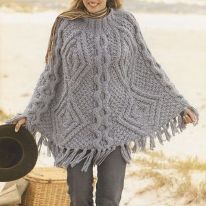 May include: A light blue knitted poncho with a cable knit pattern and fringe trim. The poncho is worn by a person with blonde hair and a scarf around their neck.