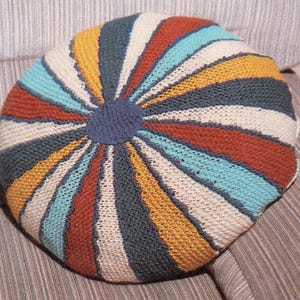 May include: A round, knitted pillow with a sunburst design. The pillow features alternating stripes of blue, teal, white, yellow, orange, and dark green radiating from a central blue circle. The pillow is placed on a striped surface.