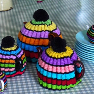 Pode incluir: Quatro capas de chá de malha coloridas em um padrão de arco-íris. As capas são feitas com listras de diferentes cores e têm pompons pretos no topo.