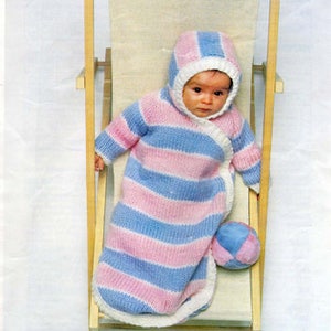 Könnte beinhalten: Ein gestrickter Schlafsack für Babys in rosa und blauen Streifen mit passender Kapuze. Der Schlafsack liegt auf einem Holzstuhl mit einem weißen Kissen. Das Baby trägt den Schlafsack und hält einen blauen und weißen Ball.