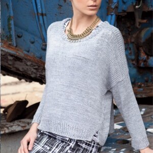May include: A light gray knit sweater with long sleeves and a round neckline. The sweater has a ribbed hem and cuffs. The model is wearing a gold-toned necklace and a black and white plaid skirt. The background is a weathered blue surface.