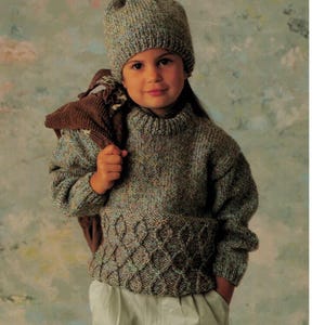 以下が含まれることがあります： ポンポンが付いた緑と茶色のニット帽をかぶった子供が、裾にケーブルニットの模様が入った緑と茶色のセーターを着ています。