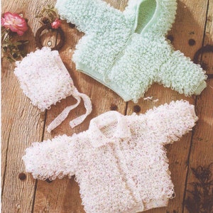 May include: A light green hooded baby jacket and a white jacket with a collar and button closure, both with a textured, looped design. A matching white bonnet with tie strings is also present. The items are displayed on a wooden surface.