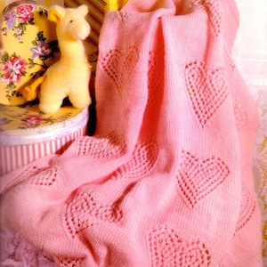 May include: A pink knitted baby blanket with a heart pattern. The blanket is folded and draped over a box with a striped pattern.