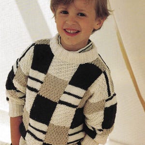 May include: A young boy wearing a white, black and beige checked jumper. The jumper has a crew neck and long sleeves.